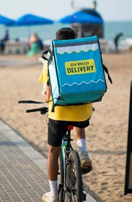 Курьер Sea Breeze Delivery с бирюзовым термокоробом едет на велосипеде вдоль пляжа