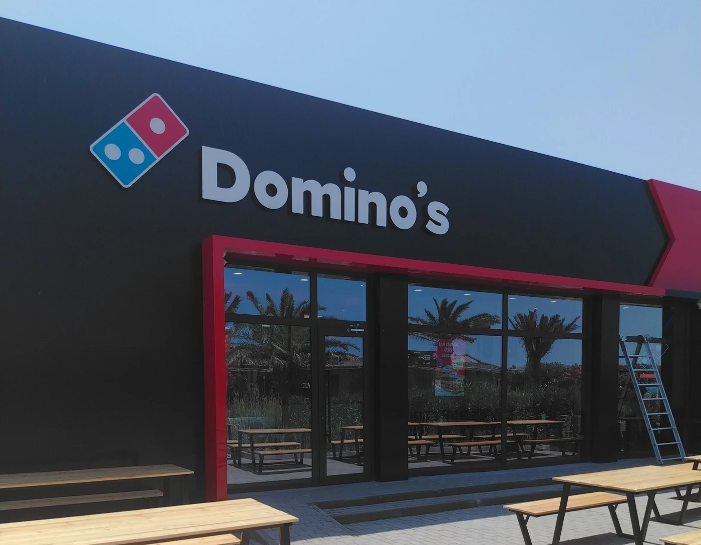 Фасад Domino's в Sea Breeze: чёрная стена с красным порталом входа, светящаяся костяшка-логотип и деревянные столы-пикники перед входом